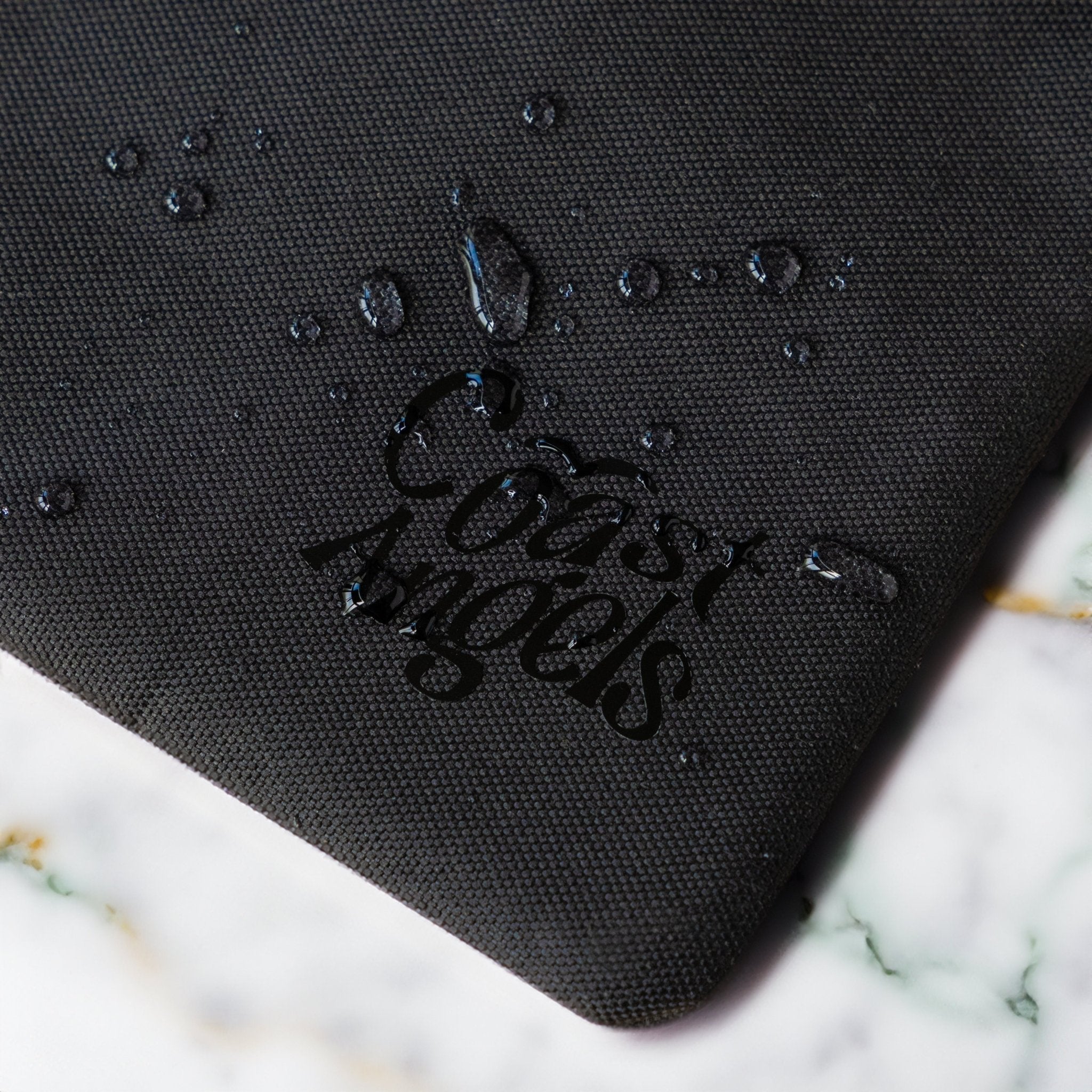 Close-up of the Coast Angels logo on a textured Soho black pouch, with water droplets beading on the surface — showcasing the bag’s water-resistant material and durable finish.