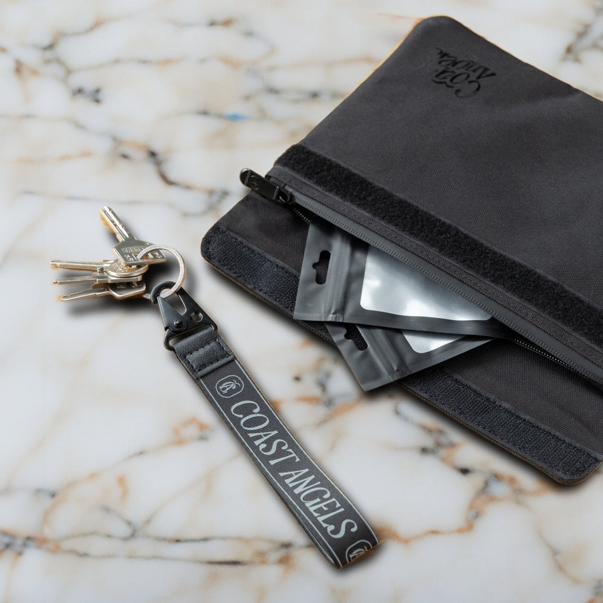 Black Coast Angels Soho smell-proof pouch shown partially unzipped on a marble surface, with resealable bags visible inside and a branded wrist strap holding a set of keys — showcasing portable, discreet functionality.